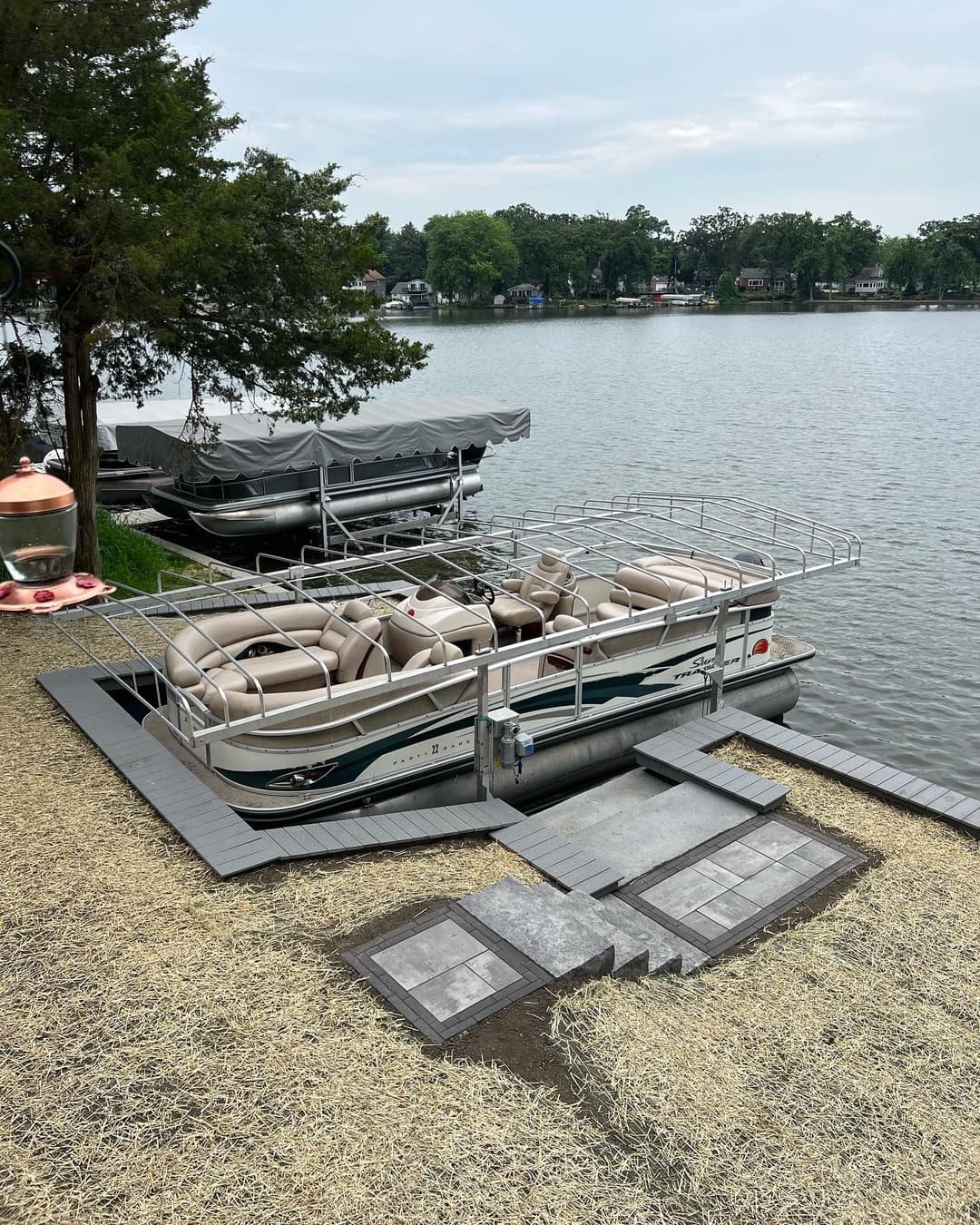 Pontoon boat docked by the lake, with another boat and tree line in the background.