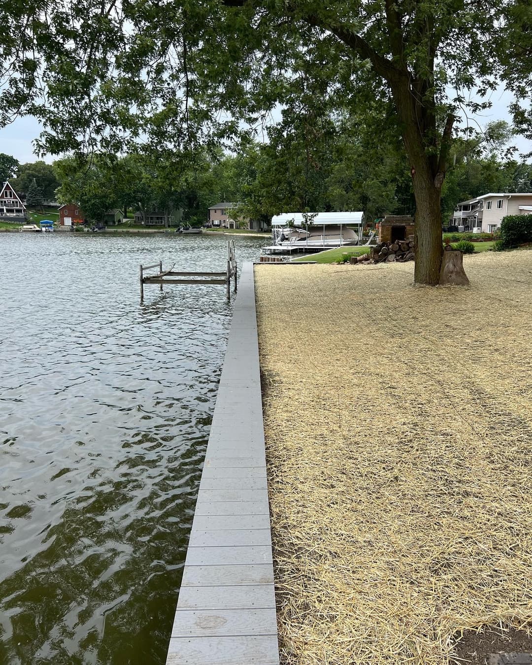Lakefront view with wooden dock, sandy shore, and green trees in a residential area.