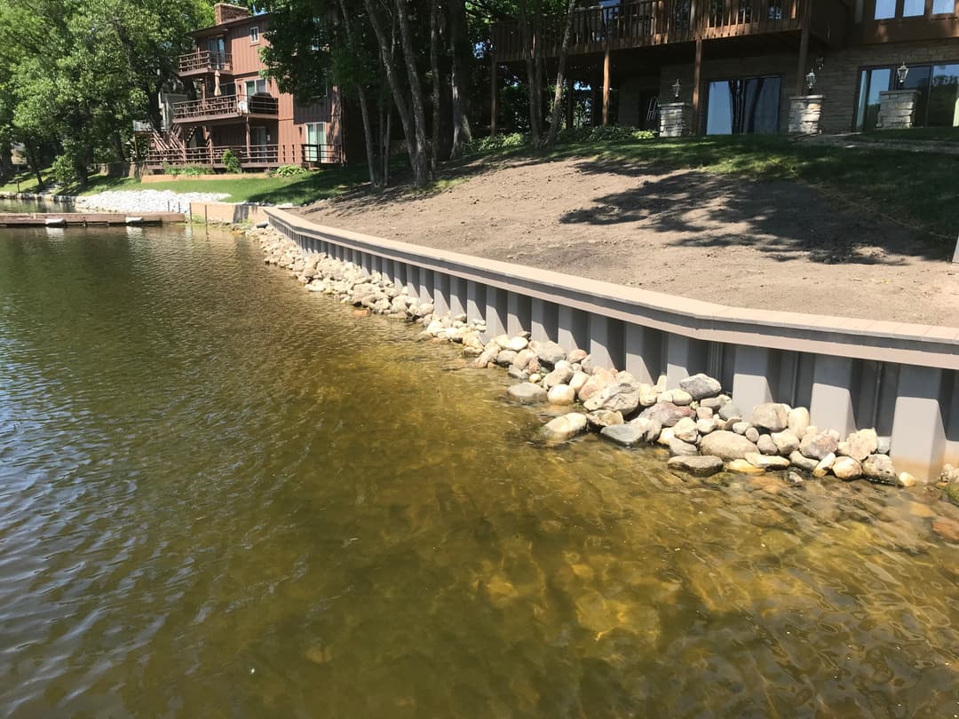 Lakefront view showing a stone-lined shoreline with homes and trees in the background.