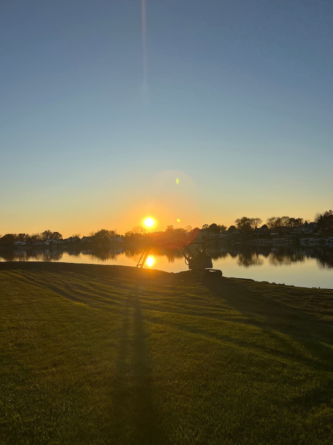 Sunset reflecting on calm water, with green grass in the foreground and silhouettes of trees.