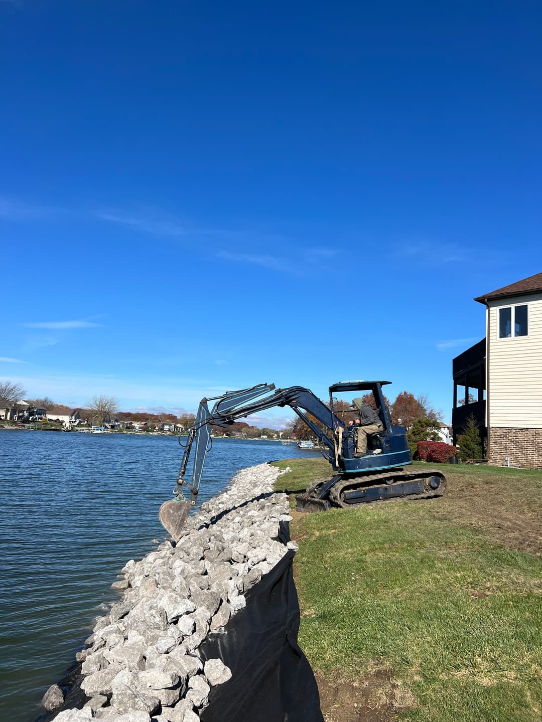 Excavator near lake building a rock barrier under a clear blue sky.