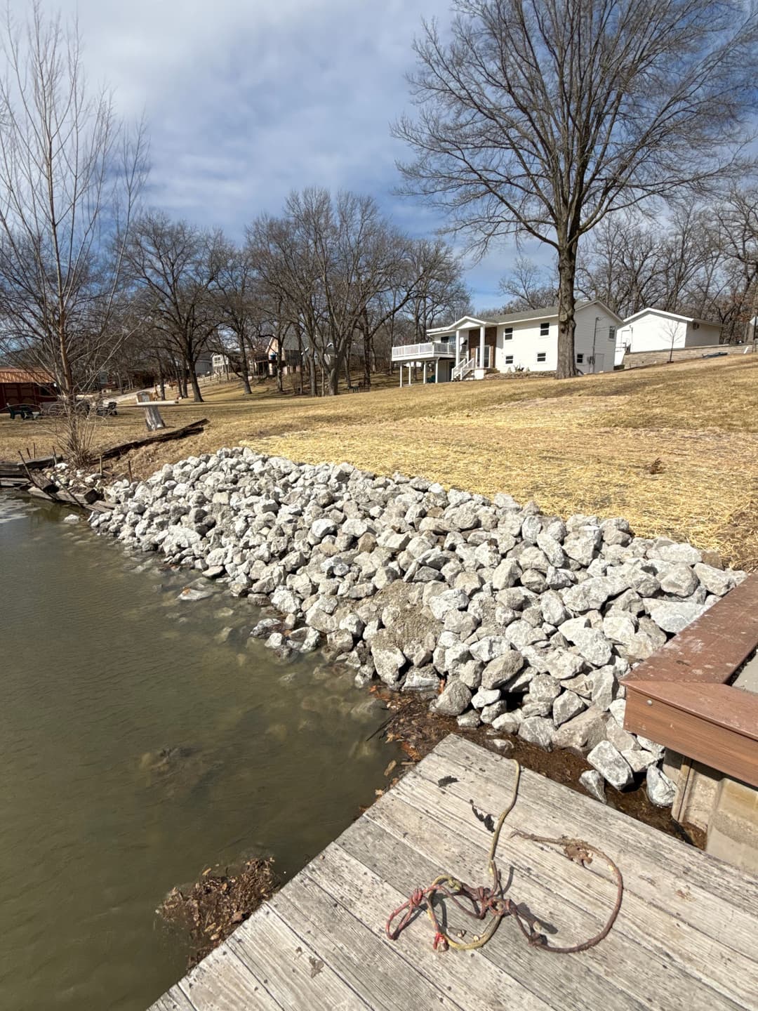 Rocky shoreline by a lake with a wooden deck and houses in the background on a cloudy day.