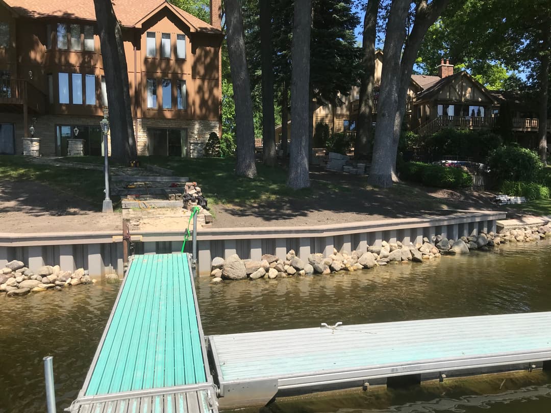Lakeside view with wooden houses, a green dock, and rocky shore under a sunny sky.