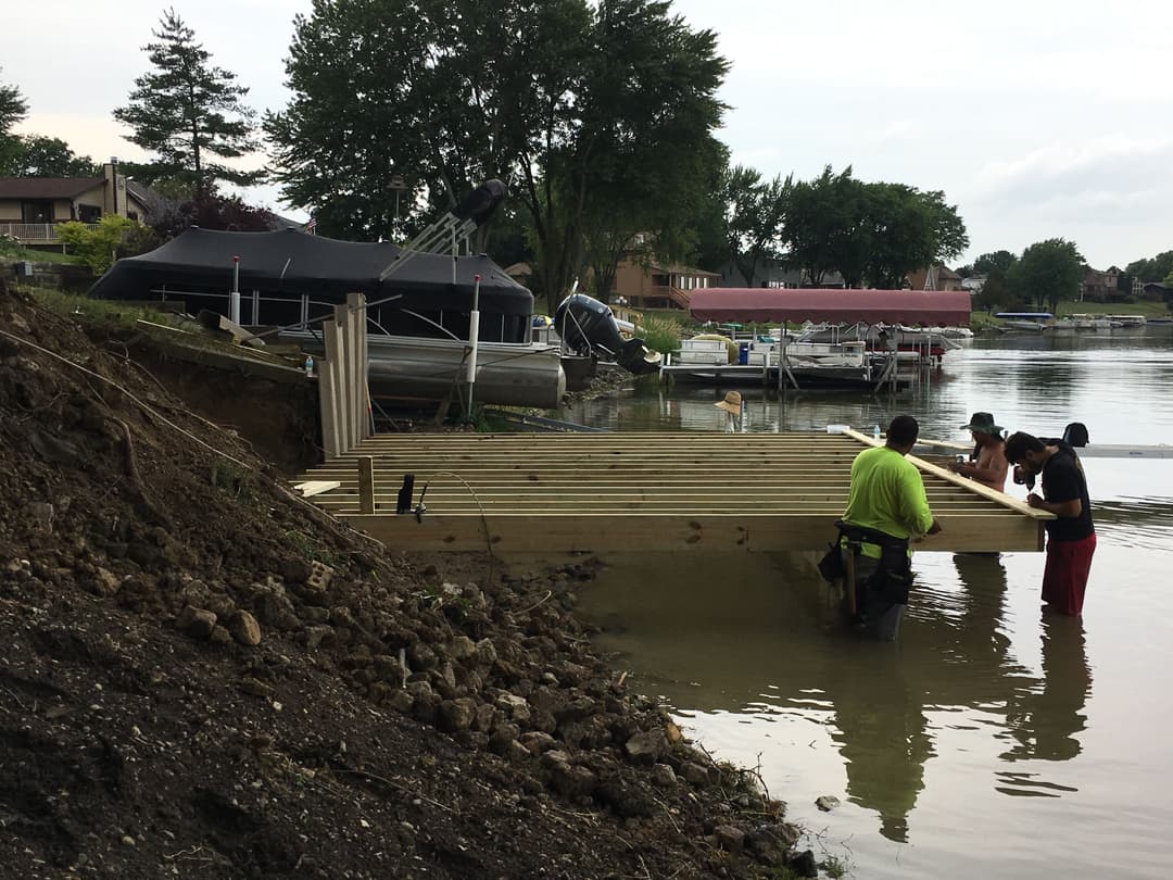 Men building a wooden dock on a tranquil lakeside, surrounded by boats and greenery.