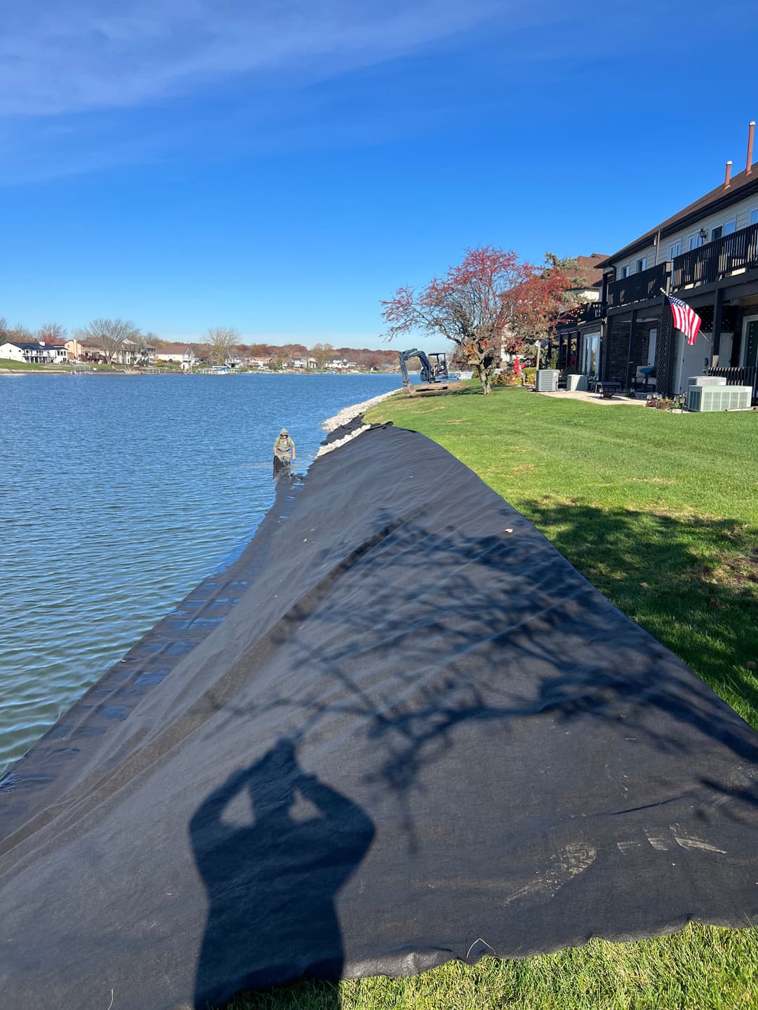 Black landscape fabric along a lake shore with blue sky, grass, and a house in background.