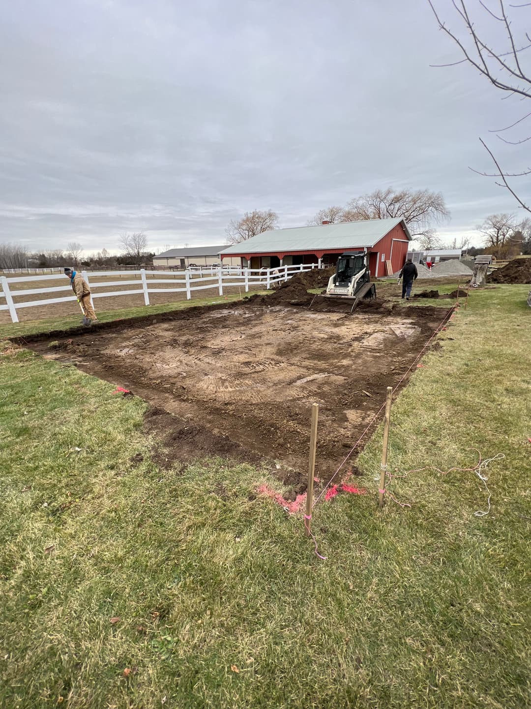 Construction site preparation with machinery on grass, near a red barn and white fence.