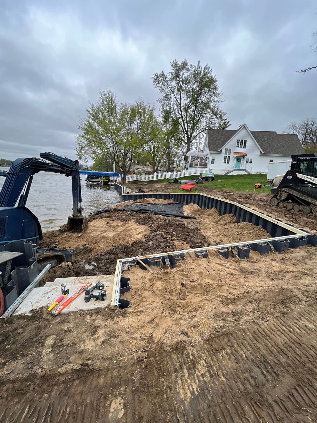 Construction of a lakeside retaining wall with machinery near a home and trees.