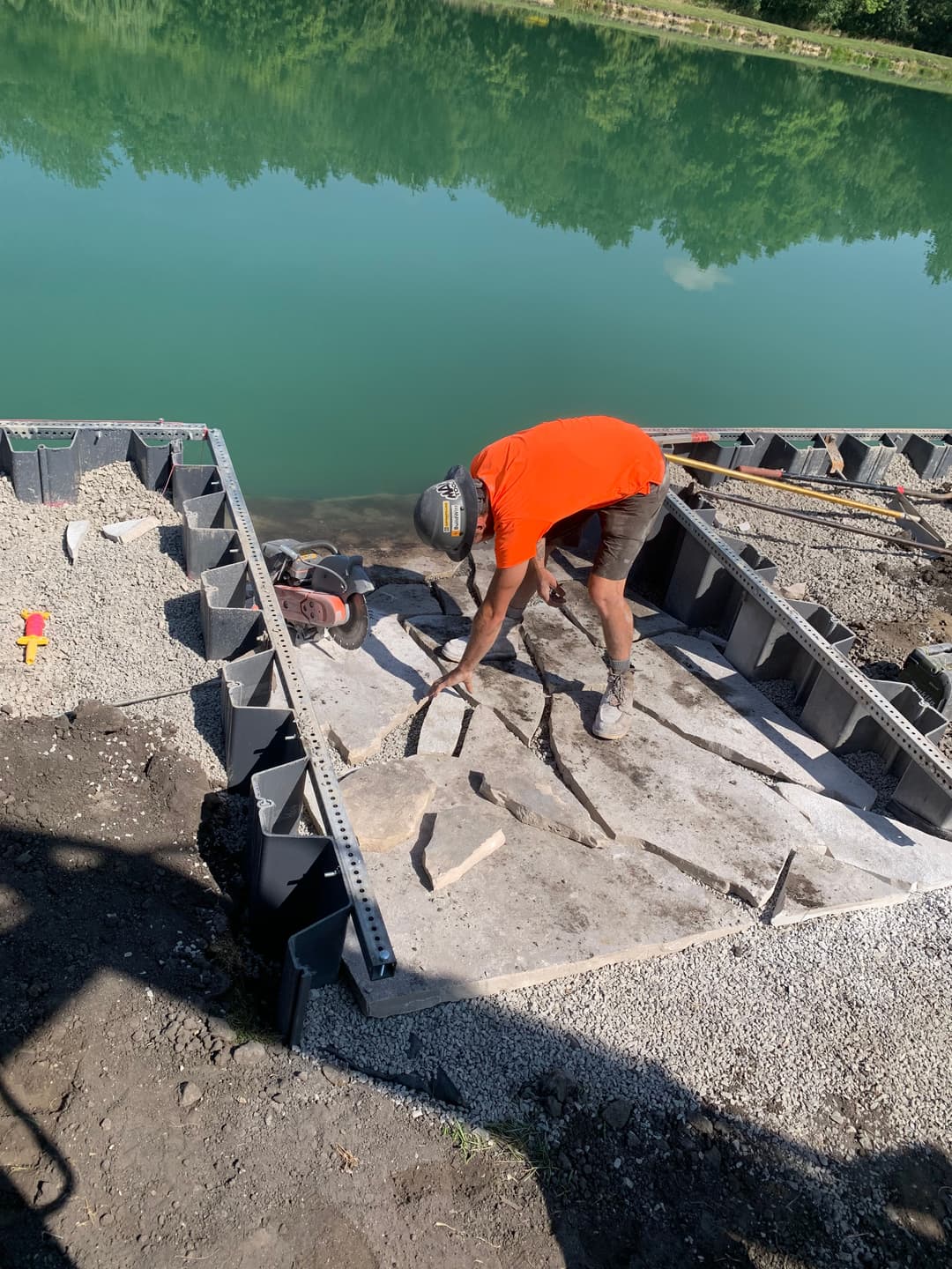 Worker laying stones on a new dock by a green lake, using construction tools and equipment.