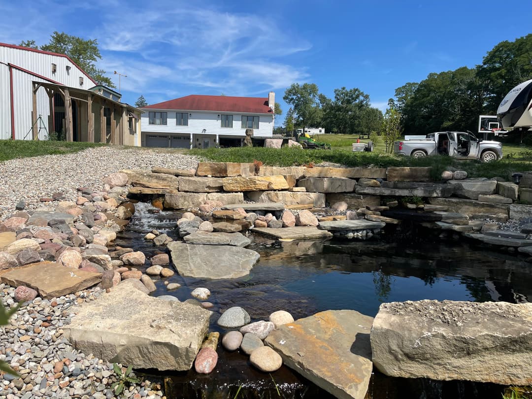 Scenic backyard pond with rocks, gravel, and a house in the background under a blue sky.