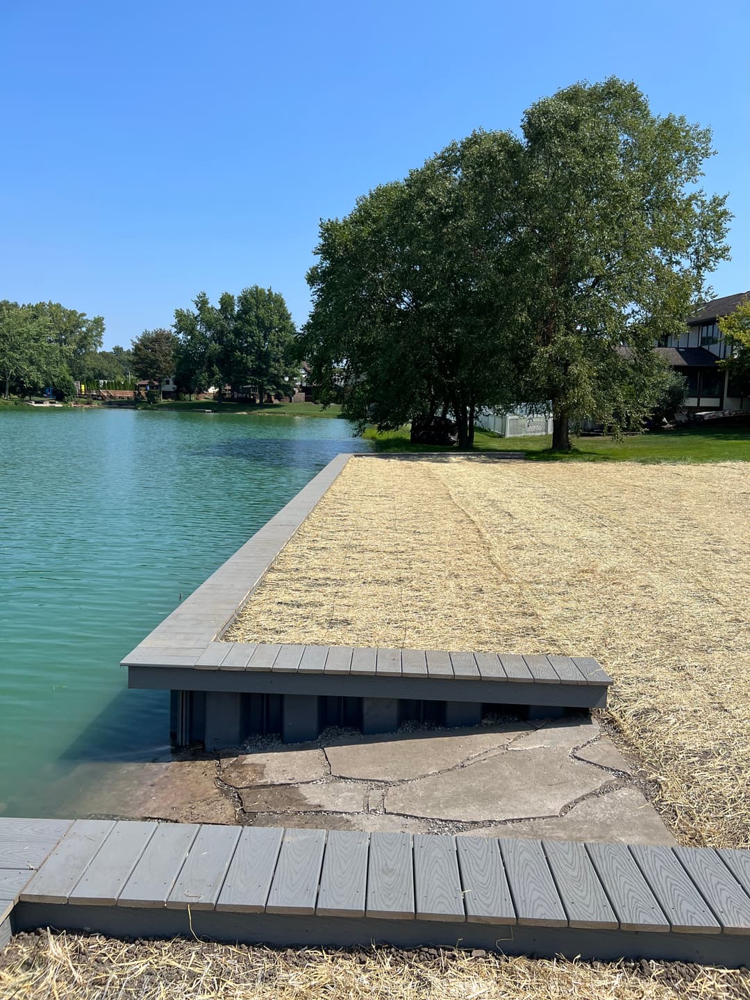 Dock extending into calm water with a grassy area and trees in the background on a sunny day.