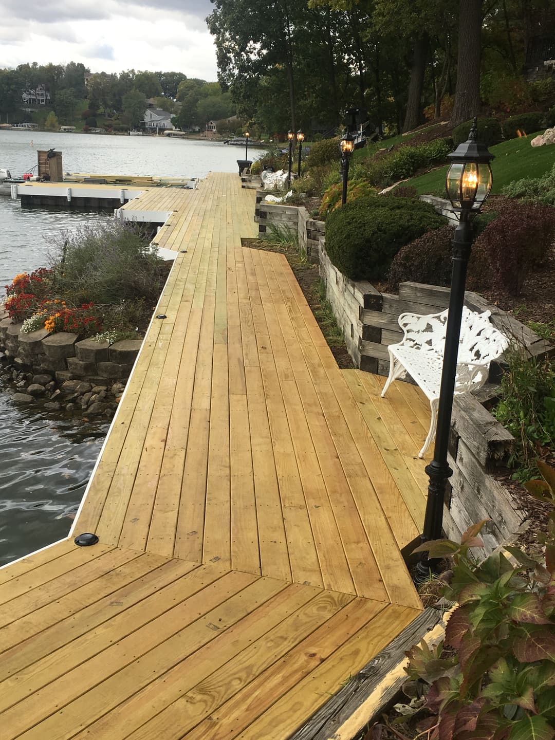 Wooden dock by a lake with lamp posts, flowers, and a white bench amidst autumn scenery.