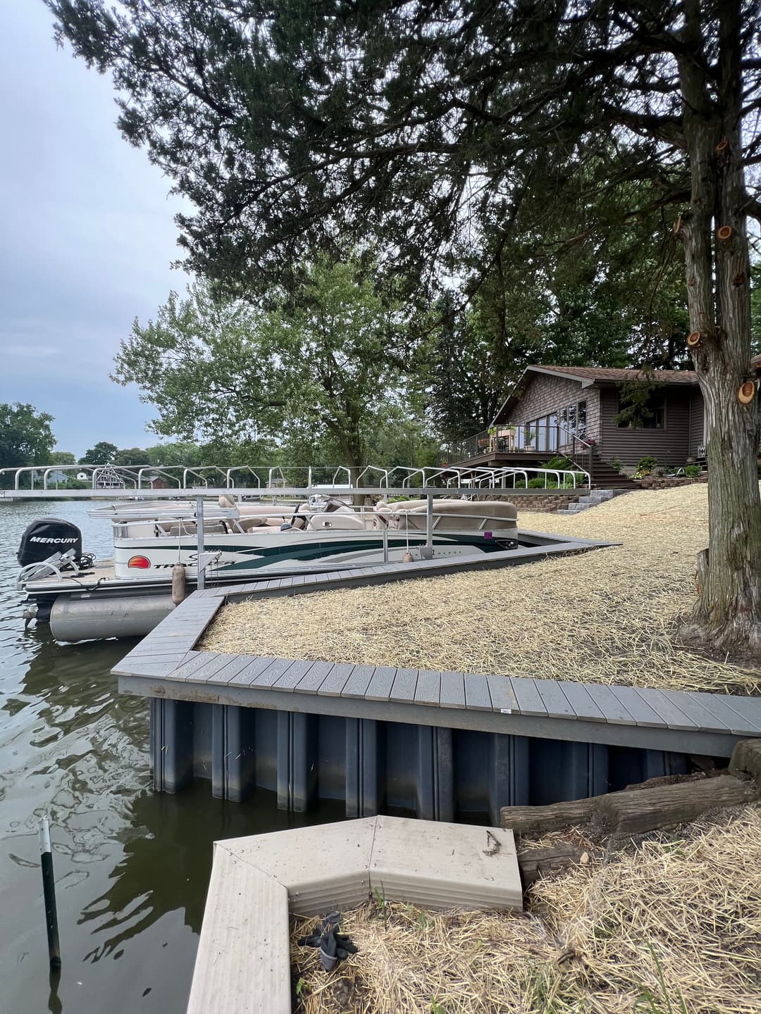 Scenic lakeside view with boats docked, wooden deck, and a house surrounded by trees.