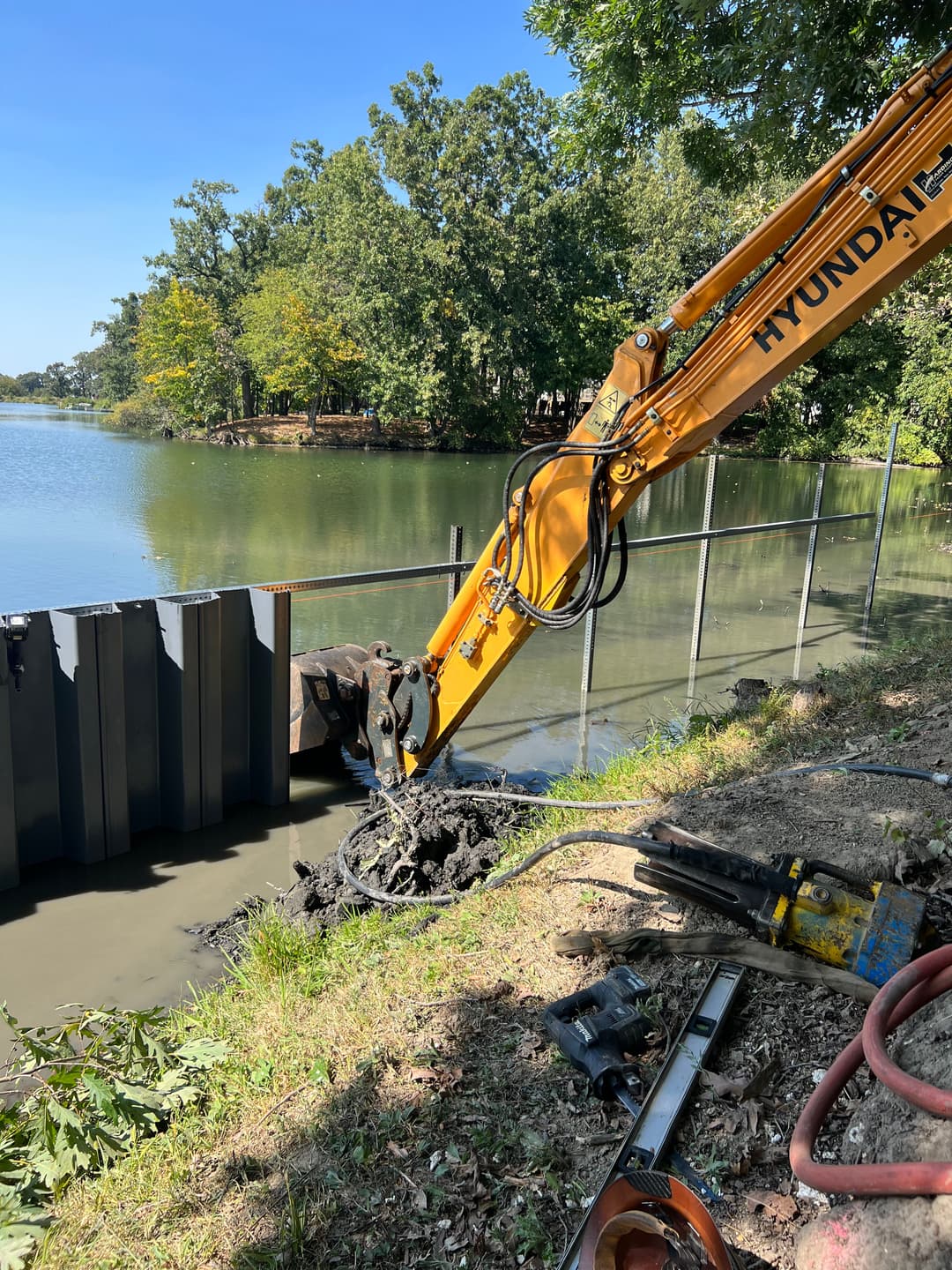 Excavator working near a lake, digging by a metal retaining wall on a sunny day.