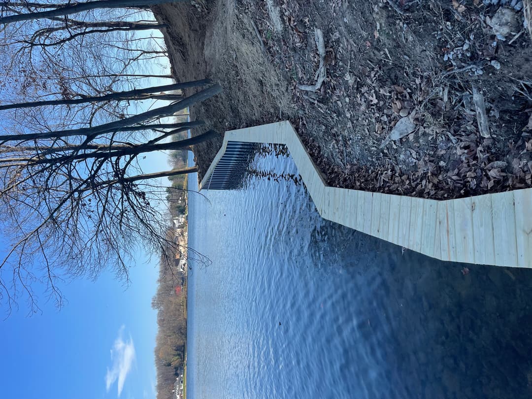Wooden boardwalk winding alongside a serene lake with trees and clear blue sky.