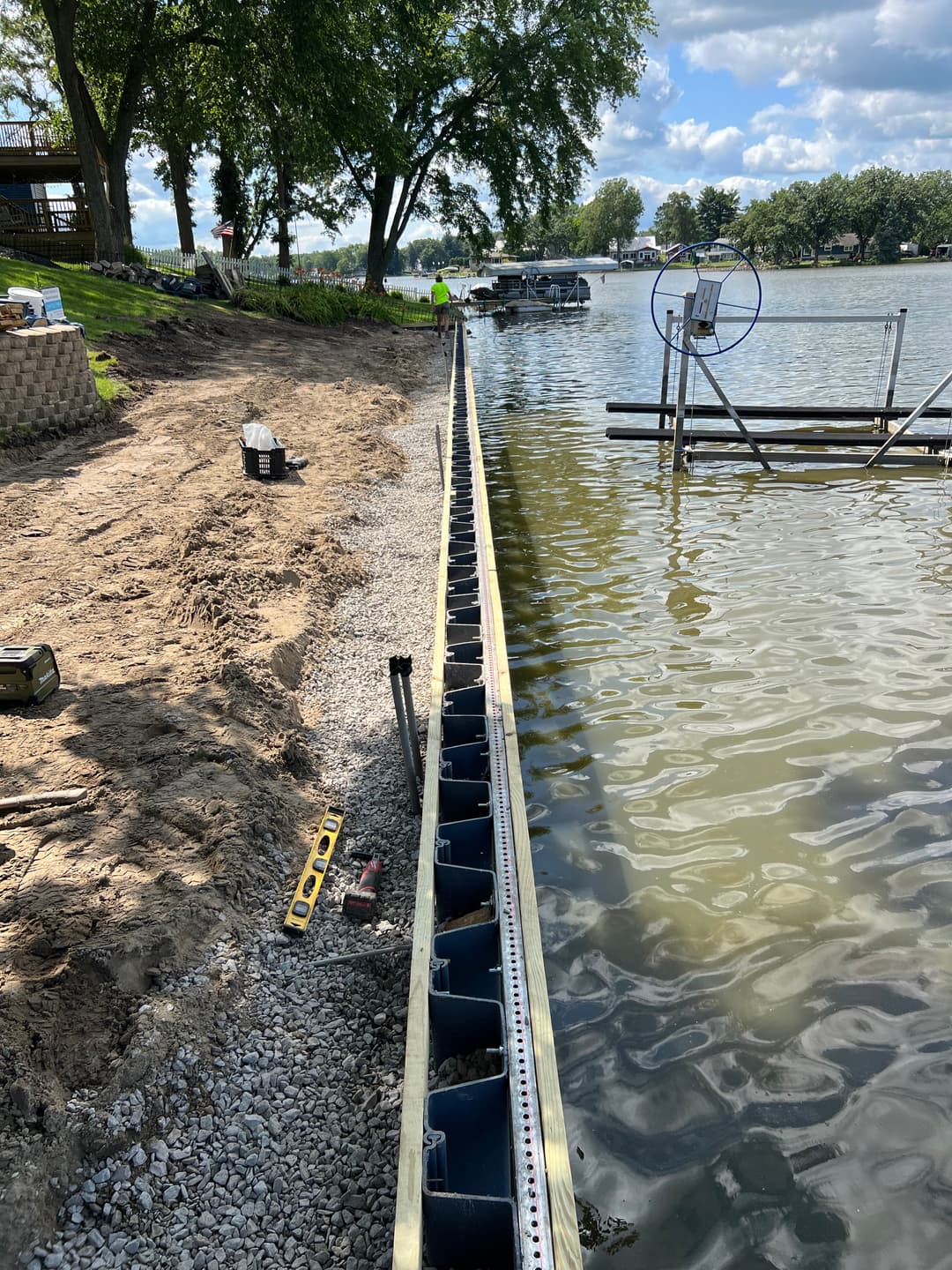 Lakefront dock construction with a wooden framework and tools on gravel shore under a clear sky.
