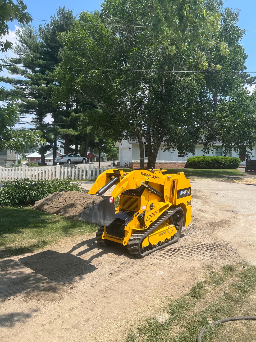 Yellow compact track loader on a dirt path surrounded by trees and houses.