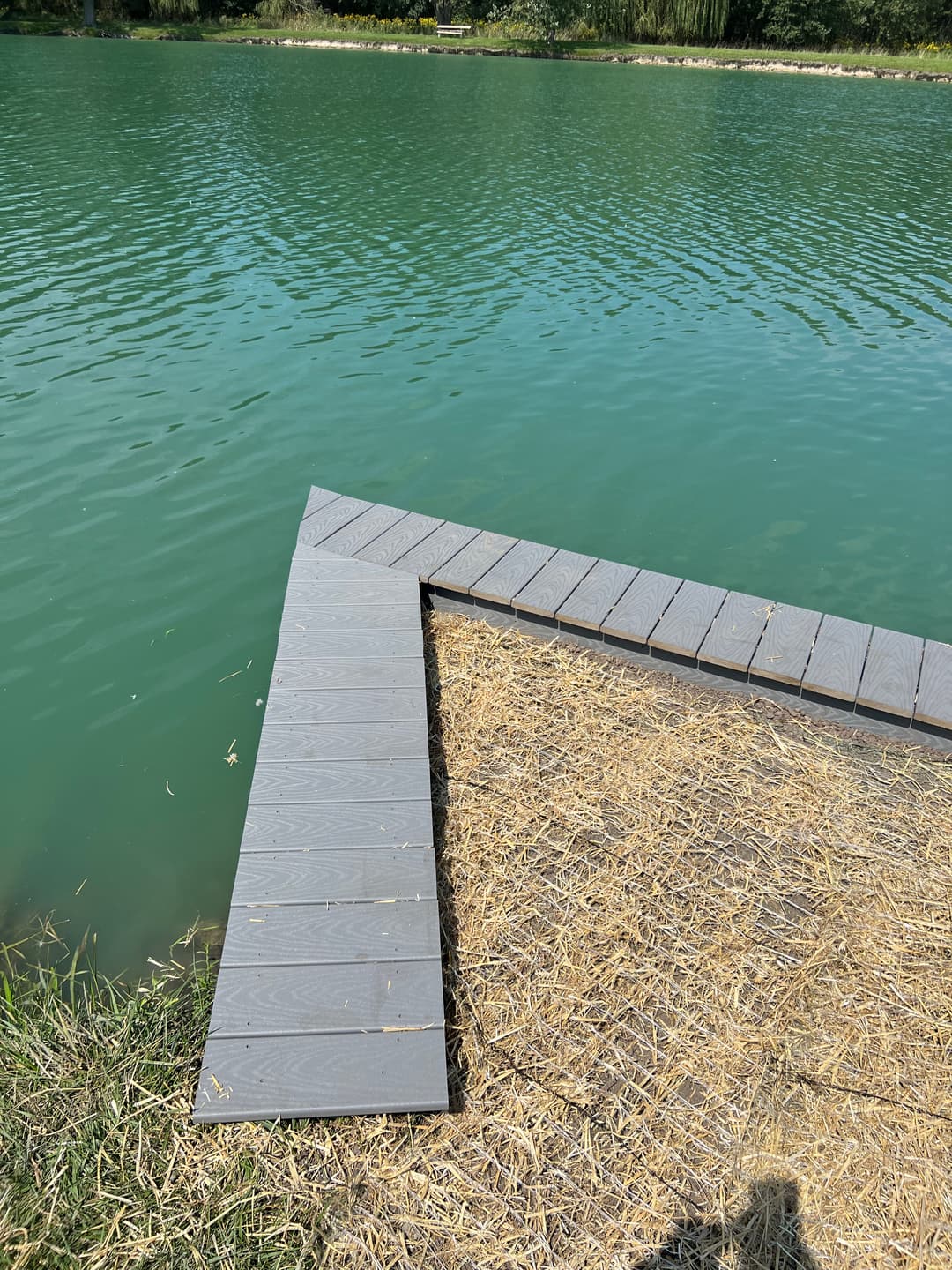Wooden dock corner extending into a turquoise lake with grassy edges and straw.