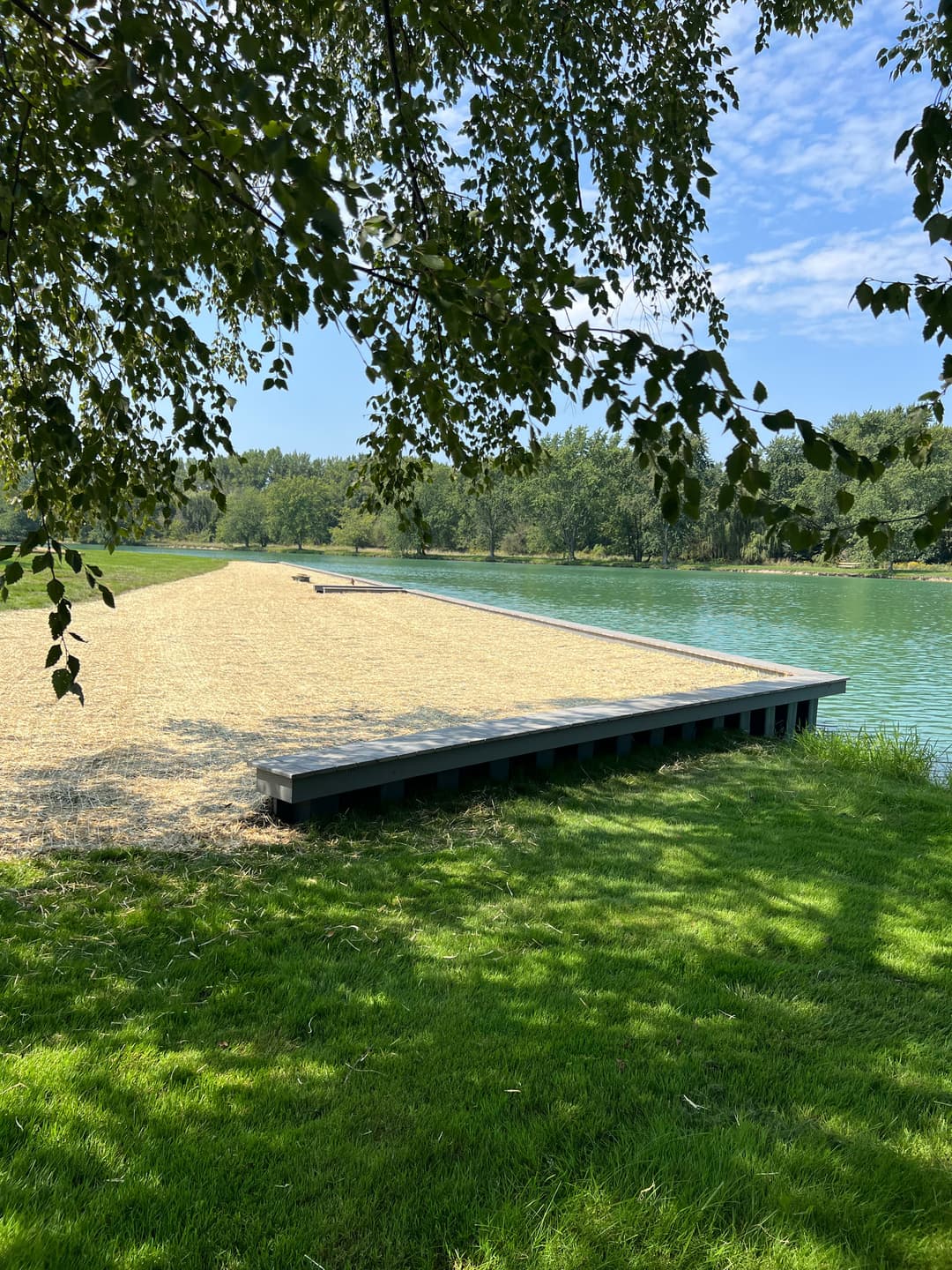 Scenic dock on a green lake surrounded by lush trees and blue sky.