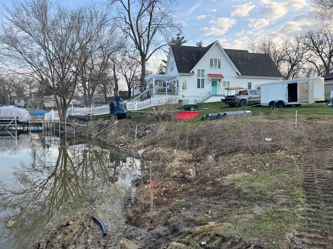 Historic white house by the water with boats, surrounded by trees and equipment.