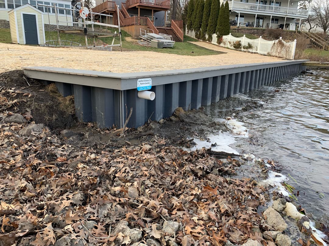 Dock extending over water with a drainage pipe, surrounded by fallen leaves and shoreline.