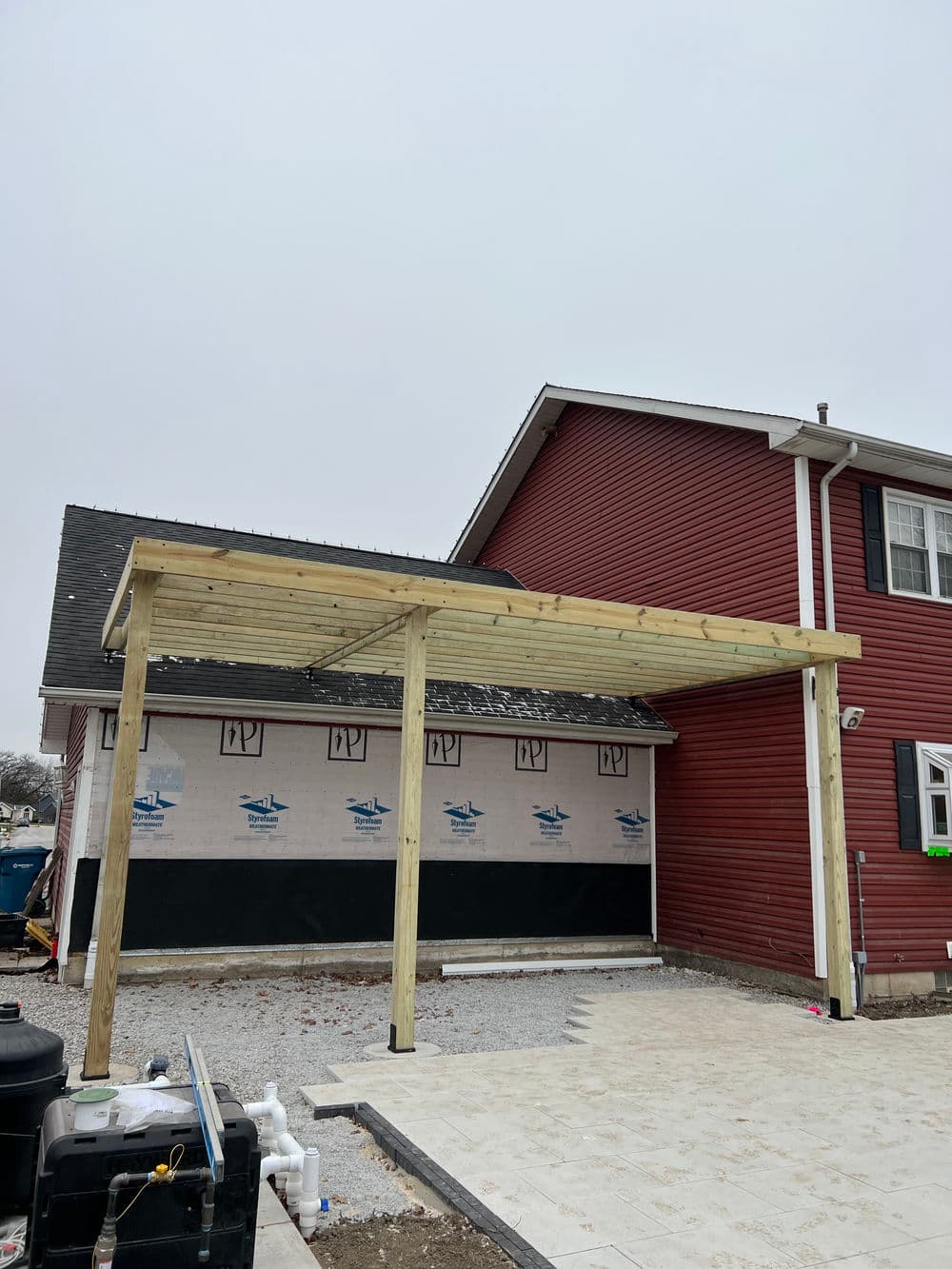 Wooden carport structure under construction next to a red house, with a gravel driveway.