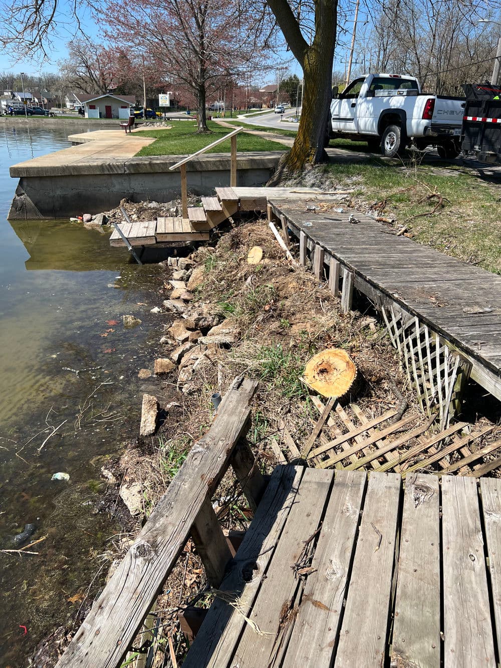 Damaged wooden dock by a lake with exposed soil, debris, and cut tree stumps in spring.