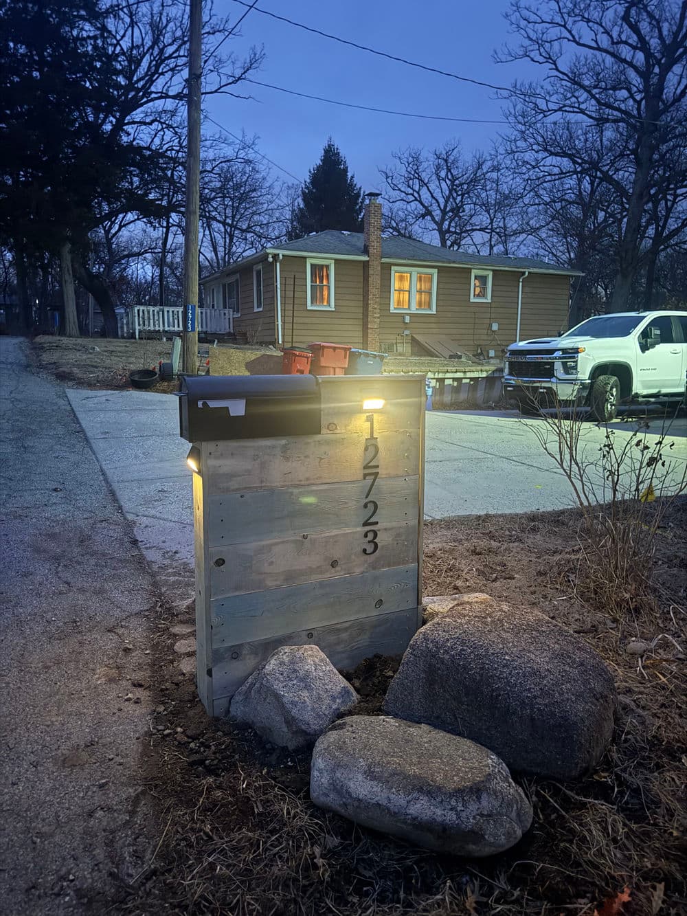Illuminated modern mailbox with the number 12723, beside a gravel driveway at dusk.