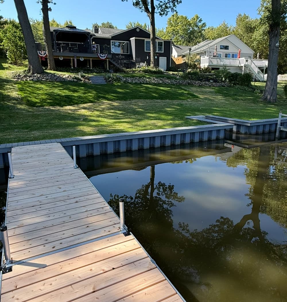 Wooden dock at a serene lakeside house with green lawn and clear water reflection.